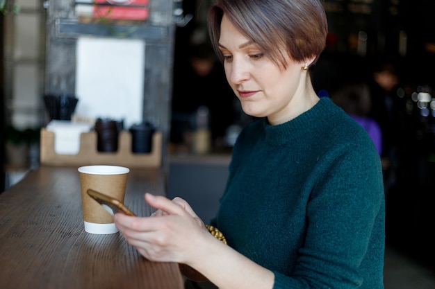 Schönes süßes Mädchen im Café mit Kaffeelächeln in die Kamera schaut. Grüner Strickpullover. modisch gekleidetes Mädchen. Mädchen schaut auf das Telefon. Geschäftsfrau