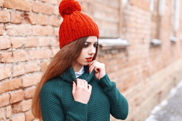 Schönes süßes Mädchen in einer roten Kappe und in der grünen Strickjacke, die nahe roter Backsteinmauer stehen