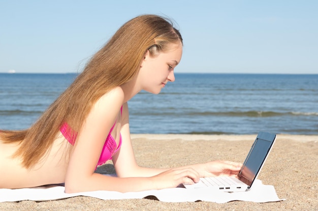 Schönes Teenager-Mädchen, das am Strand an einem weißen Laptop arbeitet