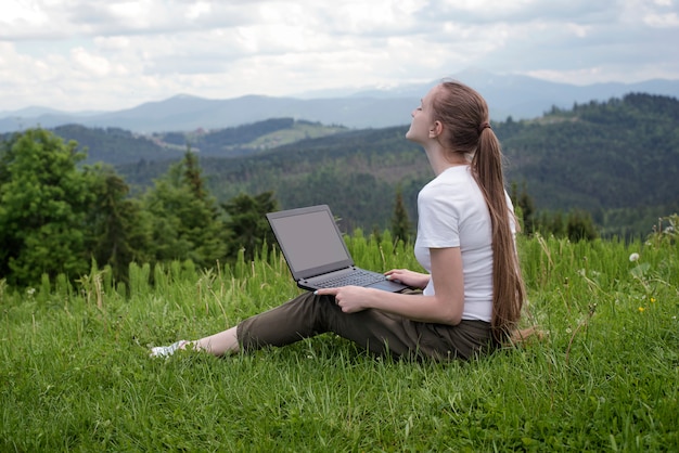 Schönes träumendes Mädchen mit einem Laptop, der auf grünem Gras auf Berge sitzt