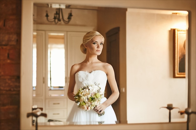 Schönes und modisches blondes Modellmädchen mit Hochzeitsfrisur im trendigen Kleid mit Blumenstrauß in ihren Händen, Hochzeitsvorbereitung der jungen Braut am Morgen