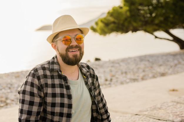 Schönes und selbstbewusstes Outdoor-Porträt eines lächelnden Mannes mit Hut und Sonnenbrille am Strand