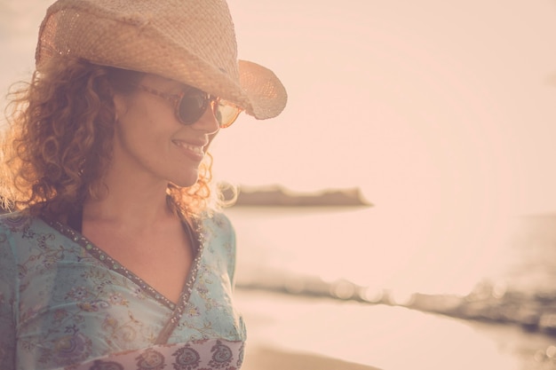 Schönes und wundervolles blondes lockiges Haarmodell mittleren Alters mit Cowboyhutlächeln und Pose am Strand mit goldenem Sonnenuntergang im Hintergrund. farbiges und freiheitsunabhängiges starkes frauenkonzept