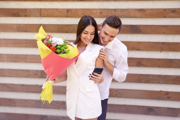 Schönes verliebtes Paar in weißen Kleidern mit einem großen Blumenstrauß, der draußen auf das Telefon schaut