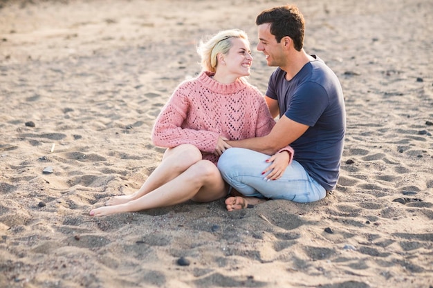 Schönes vorbildliches Paar, kaukasische junge Dame und Mann bleiben verliebt, umarmen und sitzen am Strand blonde und schwarze Haare in Beziehung Freizeitaktivitäten im Freien