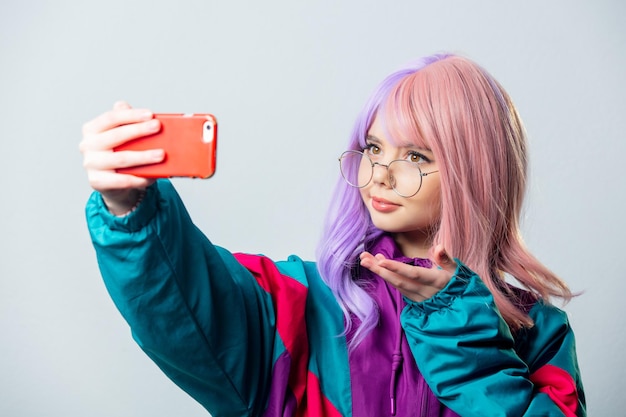 Schönes Yandere-Mädchen mit lila Haaren und 80er-Jahre-Trainingsanzug macht ein Selfie auf grauem Hintergrund