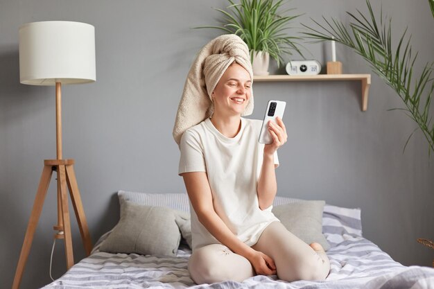 Schönheit am Morgen Gesunde, entspannte Frau, die mit dem Smartphone auf dem Bett sitzt und sich nach dem Badezimmer ausruht und einen Videoanruf hat, der lacht und positive Emotionen ausdrückt