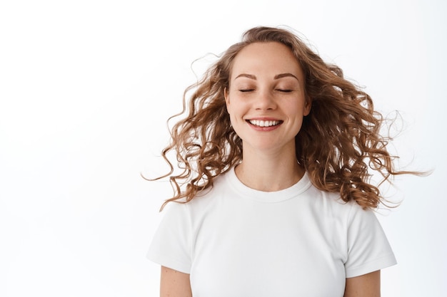 Schönheit. Blondes Mädchen schließt die Augen und genießt Wind, sanfte Brise, lockiges Haar, das in der Luft fliegt, glücklich steht und vor weißem Hintergrund lächelt