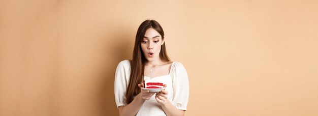 Schönheit Geburtstagskind bläst Kerze am Geburtstagskuchen, der Wunsch mit aufgeregtem Gesicht, das auf beigem Bac steht