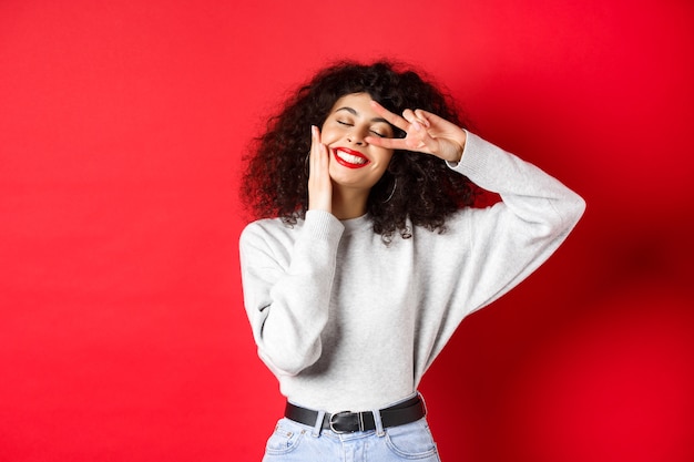 Schönheit. Hübsche Dame mit lockigem Haar und roten Lippen, Gesicht mit Make-up berührend und V-Zeichen auf Auge zeigend, sorglos lächelnd, gegen Studiohintergrund stehend.