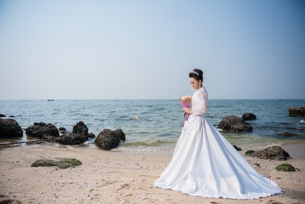 Schönheit im Hochzeitskleid, welches die Blume steht auf einem Strand hält.