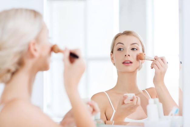 Schönheit, Make-up, Kosmetik, Morgen- und Personenkonzept - lächelnde Make-up-Pinsel und Pudergrundierung der jungen Frau, die das Badezimmer zu Hause spiegeln