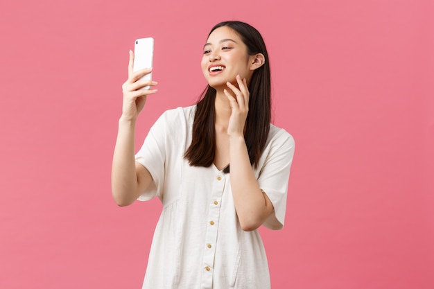 Schönheit, Menschen Emotionen und Technologiekonzept. Feminine gut aussehende stilvolle asiatische Bloggerin, die Selfie auf Smartphone-Kamera nimmt, glücklich am Handy lächelt, rosa Wand stehend