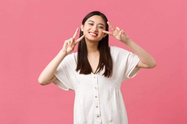Schönheit, Menschengefühle und Sommerfreizeitkonzept. Dummes und zärtliches Lächeln, glückliches japanisches Mädchen im weißen Kleid, das kawaii Friedenszeichen zeigt und freudig grinst, stehend rosa Hintergrund
