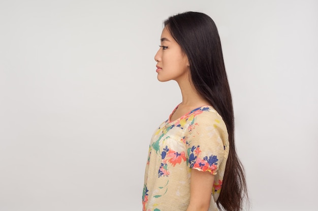 Schönheit und Hautpflege Seitenansicht eines attraktiven asiatischen Mädchens mit schönen langen brünetten Haaren in Bluse, das den Kopierraum mit ernsthaftem, ruhigem Ausdruck betrachtet, Studioaufnahme isoliert auf grauem Hintergrund