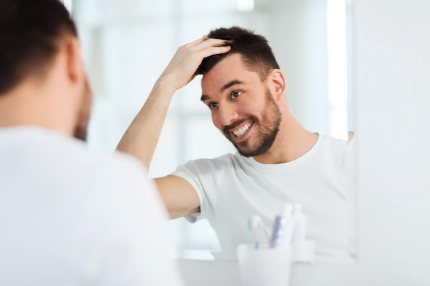 Schönheits-, Hygiene-, Frisur- und Personenkonzept - lächelnder junger Mann, der zu Hause im Badezimmer Haare spiegeln und stylen möchte
