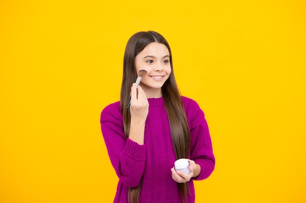 Schönheits-Make-up und Kinderkosmetik Teenager-Mädchen mit Pinsel, das Rouge auf das Gesicht aufträgt, isoliert auf Wellow-Hintergrund