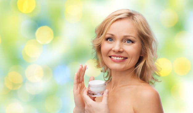 Schönheits-, Menschen-, Hautpflege- und Kosmetikkonzept - glückliche Frau, die Creme auf ihrem Gesicht über grünem Sommerlichthintergrund aufträgt