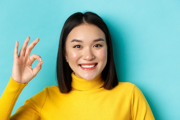 Schönheits- und Make-up-Konzept. Schließen Sie oben von der glücklichen asiatischen Frau mit der gesunden glänzenden Haut, die OK-Zeichen zeigt und zufrieden lächelt und über blauem Hintergrund steht.