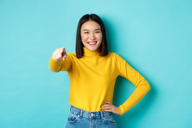 Schönheits- und Modekonzept. Fröhliche asiatische Frau, die lacht und lächelt, Finger auf Kamera zeigt, Sie wählen, über blauem Hintergrund stehend.