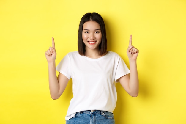 Schönheits- und Modekonzept. Schöne asiatische Frau im weißen T-Shirt, das Finger oben zeigt, über gelbem Hintergrund stehend.