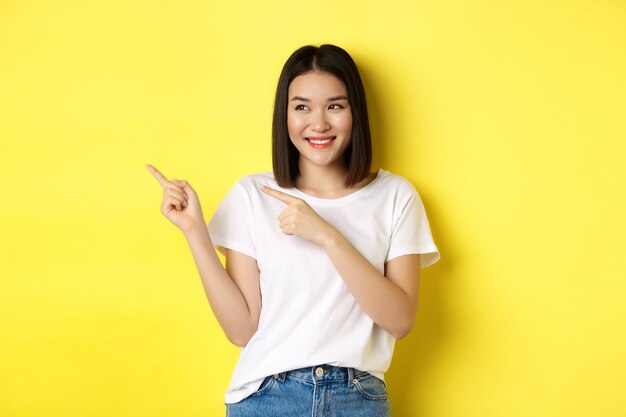 Schönheits- und Modekonzept. Schöne asiatische Frau im weißen T-Shirt zeigt Finger links, stehend über gelbem Hintergrund.