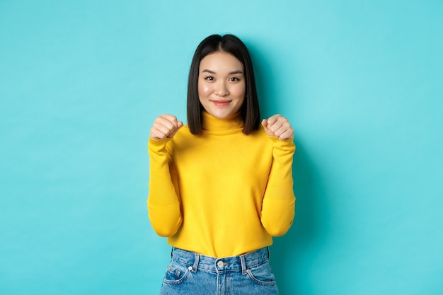 Schönheits- und Modekonzept. Schöne und stilvolle asiatische Frau im gelben Pullover, die die Hände in der Nähe der Brust hält, als ob sie ein Banner oder ein Logo halten würde, auf blauem Hintergrund stehend