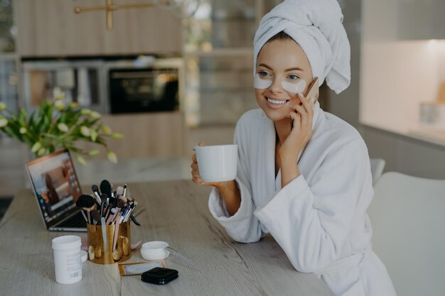 Schönheitsbehandlung Körperpflege- und Hygienekonzept Schöne junge Frau trägt kosmetische Pflaster unter den Augen auf, trägt einen Bademantel, trinkt Kaffee, spricht mit einem Freund über das Smartphone und sitzt am Tisch über dem Innenraum