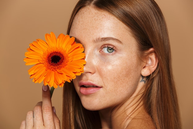 Schönheitsbild der herrlichen hemdlosen Frau 20s stehend mit orange Blume, lokalisiert über beigem Hintergrund