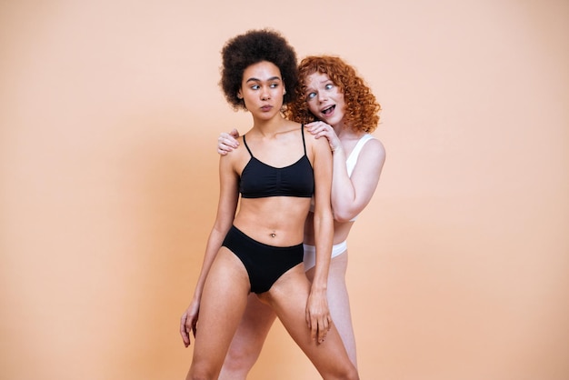 Schönheitsbild von zwei jungen Frauen mit unterschiedlicher Haut und unterschiedlichem Körper, die im Studio für ein körperpositives Fotoshooting posieren. Gemischte weibliche Models in Dessous auf farbigen Hintergründen