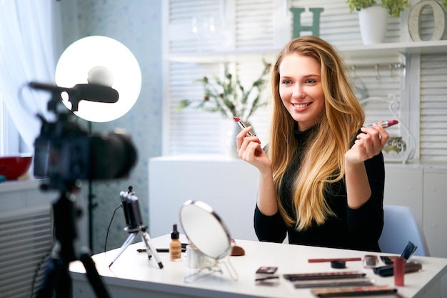 Schönheitsbloggerin filmt tägliche Make-up-Routine-Tutorial in der Nähe der Kamera auf einem Stativ Influencer blonde Mädchen Live-Streaming Kosmetik-Produktbewertung im Home-Studio Vlogger Frau, die Lippenstiele vergleicht