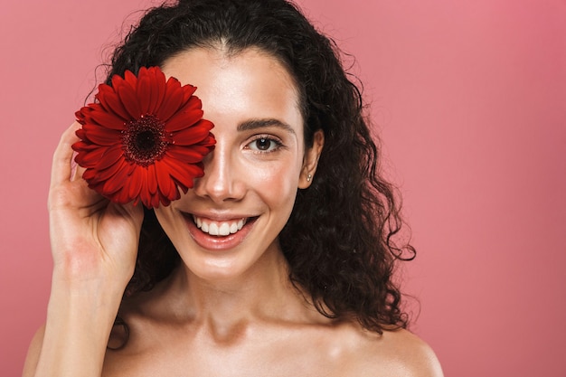 Schönheitsfoto der europäischen nackten Frau 20s mit den langen Haaren, die lächeln und rote Blume halten, lokalisiert über rosa Wand