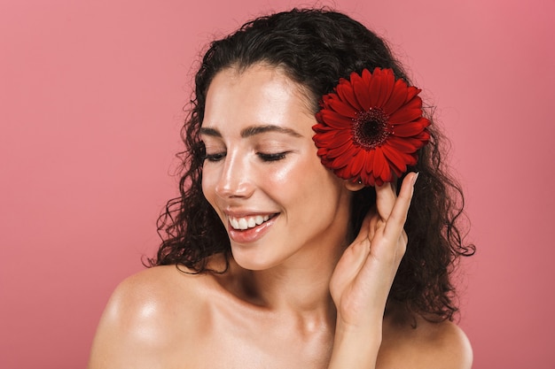 Schönheitsfoto der europäischen nackten Frau 20s mit langen Haaren und ohne Make-up, das rote Blume hält, lokalisiert über rosa Wand