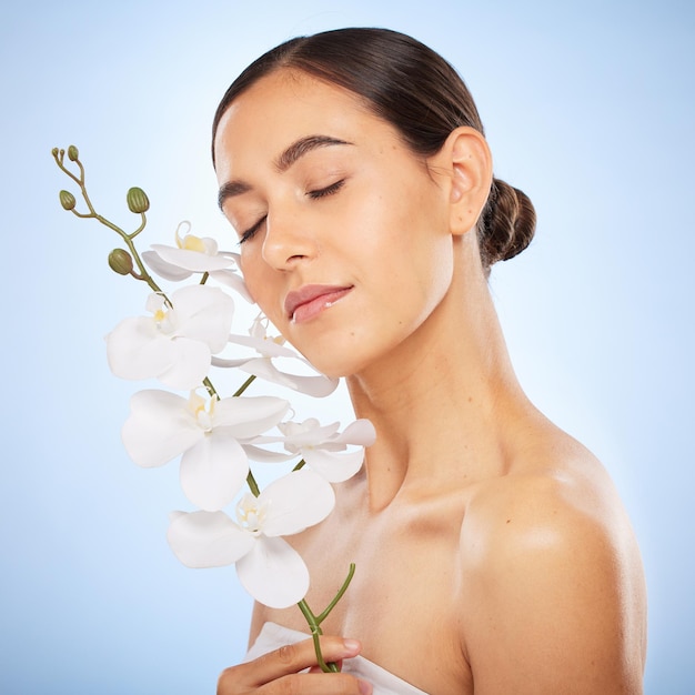 Schönheitspflege und Frau mit einer Orchidee in einem Studio für eine gesunde Wellness- und natürliche Gesichtsroutine Kosmetische Selbstpflege und Mädchenmodell mit klarer Haut von der Spa-Gesichtsbehandlung durch blauen Hintergrund