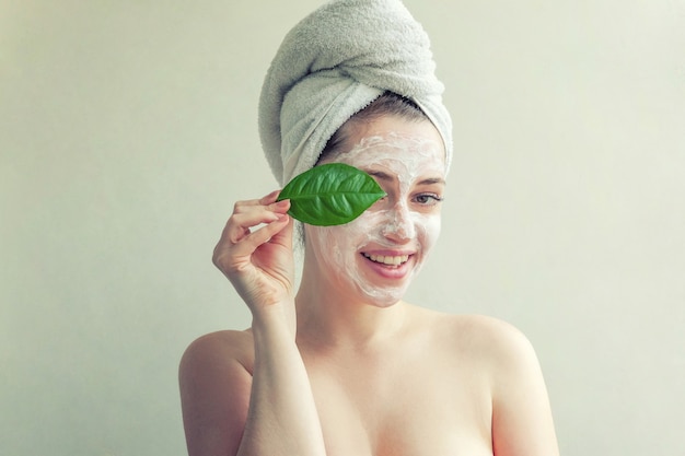 Schönheitsporträt der Frau im Handtuch auf dem Kopf mit weißer Nährmaske oder Creme im Gesicht und grünem Blatt in der Hand, weißer Hintergrund isoliert. Hautpflege reinigendes Öko-Bio-Kosmetik-Spa-Konzept