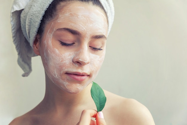 Schönheitsporträt der Frau im Handtuch auf dem Kopf mit weißer Nährmaske oder Creme im Gesicht und grünem Blatt in der Hand, weißer Hintergrund isoliert. Hautpflege reinigendes Öko-Bio-Kosmetik-Spa-Konzept