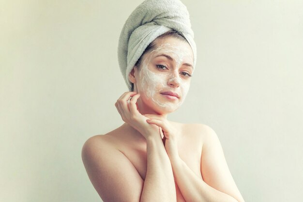 Schönheitsporträt der Frau im Handtuch auf dem Kopf mit weißer pflegender Maske oder Creme im Gesicht, weißer Hintergrund isoliert. Hautpflege reinigendes Öko-Bio-Kosmetik-Spa-Relax-Konzept