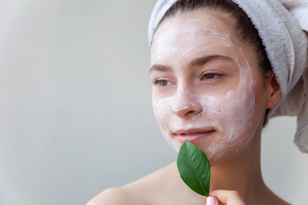 Schönheitsporträt der Frau im Handtuch auf Kopf mit weißer pflegender Maske oder Creme auf Gesicht und grünem Blatt in der Hand