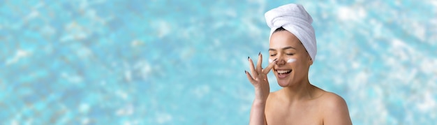 Schönheitsporträt der Frau im weißen Handtuch auf dem Kopf trägt Creme auf das Gesicht auf. Hautpflege-Reinigung Öko-Bio-Kosmetik-Spa-Relax-Konzept.