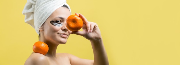 Schönheitsporträt der frau in weißem handtuch auf dem kopf mit goldener pflegemaske im gesicht hautpflege-reinigung öko-bio-kosmetik-spa-relax-konzept ein mädchen steht mit dem rücken und hält eine orangefarbene mandarine