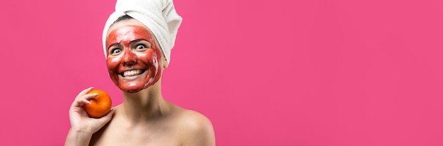 Schönheitsporträt der Frau in weißem Handtuch auf dem Kopf mit roter pflegender Maske im Gesicht Hautpflege-Reinigung Öko-Bio-Kosmetik-Spa-Relax-Konzept Ein Mädchen steht mit dem Rücken und hält eine orangefarbene Mandarine