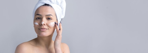 Schönheitsporträt der Frau in weißem Handtuch auf dem Kopf trägt Creme auf das Gesicht auf Hautpflege-Reinigung Öko-Bio-Kosmetik-Spa-Relax-Konzept