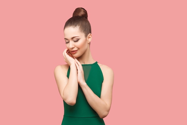 Schönheitsporträt der glücklichen schönen jungen Frau mit Brötchenfrisur und Make-up im grünen Kleid, das mit geschlossenen Augen steht, ihr Gesicht berührt und lächelt. Indoor-Studioaufnahme, isoliert auf rosa Hintergrund.