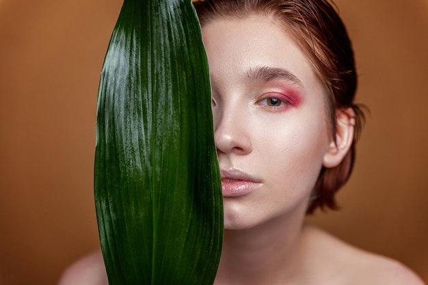 Schönheitsporträt der jungen brünetten Frau mit perfektem Hautmode-Make-up und grünem Blatt auf natürlichem braunem Hintergrund. Reinigendes Öko-Bio-Kosmetik-Entspannungs- und Hautpflegekonzept. Spa und Wellness.