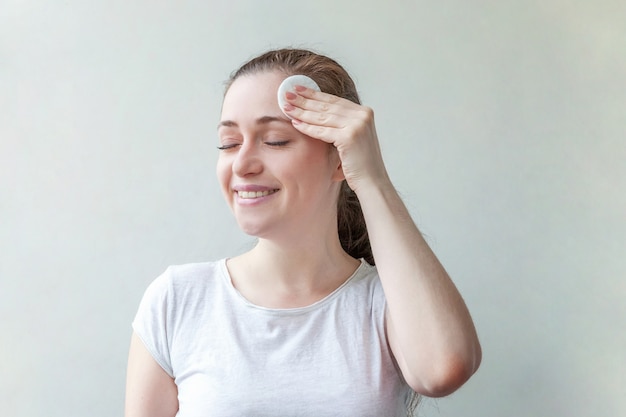 Schönheitsporträt der lächelnden Frau mit der weichen gesunden Haut, die Make-up mit Wattepad entfernt auf weißem Hintergrund entfernt.