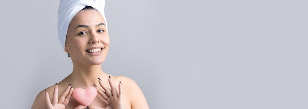 Schönheitsporträt einer Frau in weißem Handtuch auf dem Kopf mit einem Schwamm für einen Körper im Hinblick auf ein rosa Herz Hautpflege-Reinigung Öko-Bio-Kosmetik-Spa-Relax-Konzept