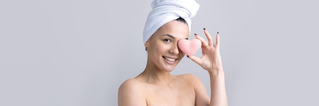 Schönheitsporträt einer Frau in weißem Handtuch auf dem Kopf mit einem Schwamm für einen Körper im Hinblick auf ein rosa Herz Hautpflege-Reinigung Öko-Bio-Kosmetik-Spa-Relax-Konzept