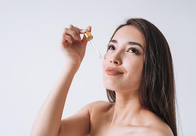 Schönheitsporträt einer glücklich lächelnden asiatischen Frau mit dunklen langen Haaren mit Pipette mit Bio-Öl auf sauberem, frischem Hautgesicht und Händen auf weißem Hintergrund isoliert