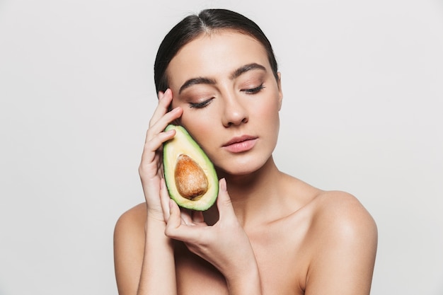 Schönheitsporträt einer jungen gesunden attraktiven brünetten Frau, die lokal steht und mit einer geschnittenen Avocado aufwirft