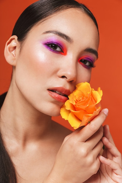 Schönheitsporträt einer jungen topless asiatischen Frau mit brünetten Haaren, die helles Make-up trägt, isoliert auf Rot stehend, mit einer gelben Rose posieren